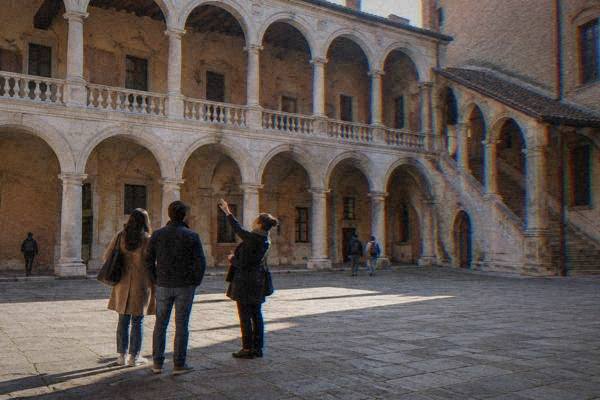 A local guide and a couple in a courtyard with two storeys of pale stone arcades, the guide gesturing up at the arches, Palazzo del Bo