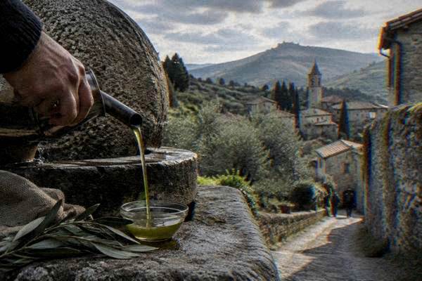 A hand pouring fresh green olive oil from a small steel spout into a small glass bowl beside a stone olive press, Arqua Petrarca