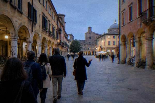 A small walking group crossing a pale paved square behind a guide, low stone arcades and ochre houses on both sides, Centro Storico