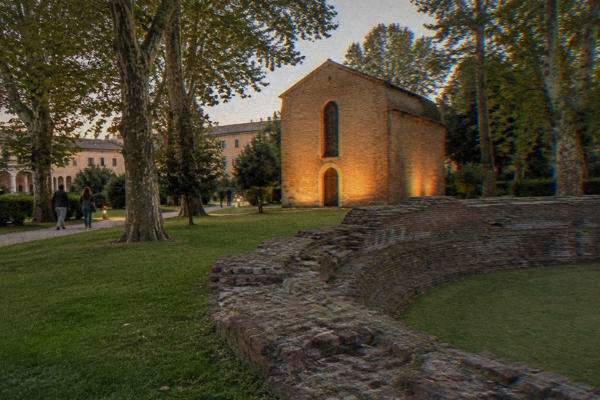 A small brick chapel glowing under warm lamplight in a green garden at dusk, a few visitors small and distant on the path, Scrovegni Chapel