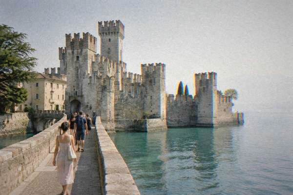 A small group of day visitors crossing a stone bridge toward a moated castle with crenellated towers on a lake peninsula, Sirmione