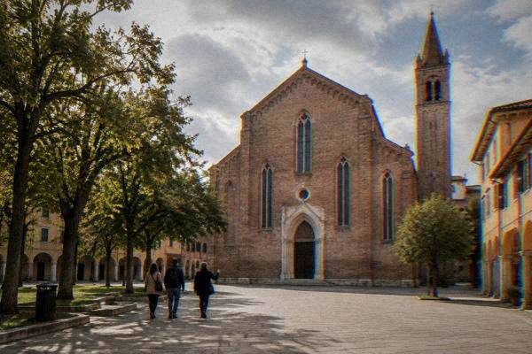 A private guide and two visitors crossing a small tree-lined square before a plain Gothic red-brick church, Chiesa degli Eremitani
