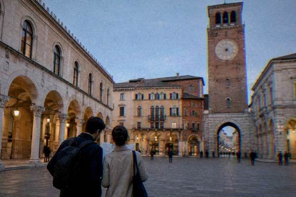 A pair of friends seen from behind studying a folded paper in a quiet square beside a clock tower, looking up at the facades, Piazza dei Signori