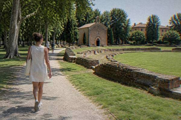 A lone visitor with earphones in, walking a gravel path in a green garden toward a small plain red-brick chapel beside low brick remains of an oval amphitheatre, Giardini dell'Arena
