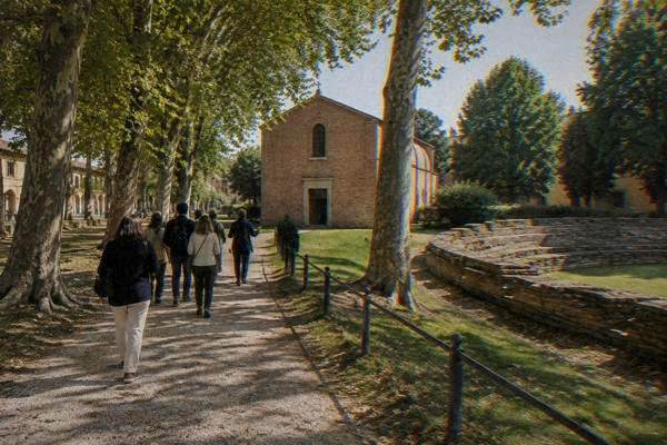 A small group walking in a loose line behind a guide along a tree-lined path toward a small plain red-brick chapel, Scrovegni Chapel