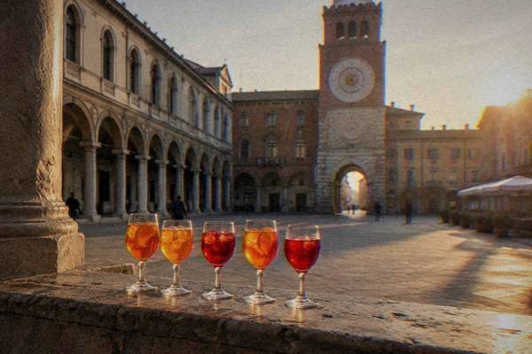 Five small glasses of orange and red aperitifs lined up on a stone ledge in a quiet square at golden hour, Piazza dei Signori