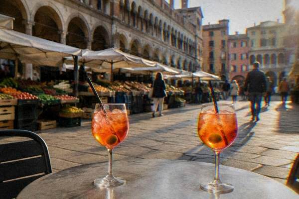 Two tall glasses of an orange aperitif with an olive on a small round table on a paved square, passers-by blurred behind, Piazza delle Erbe
