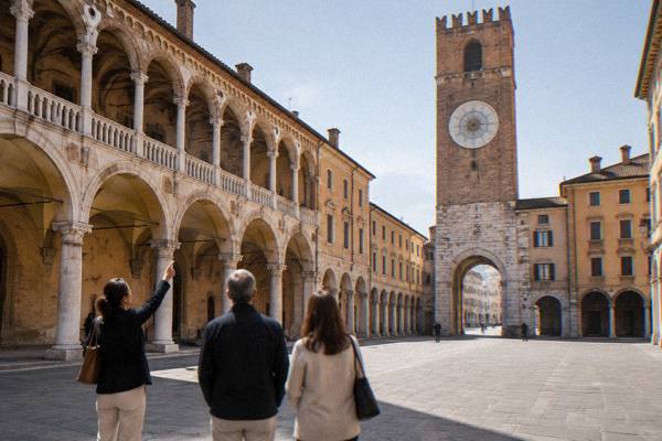A private guide pointing up at a stone tower while two visitors look on in a quiet paved square, Piazza dei Signori