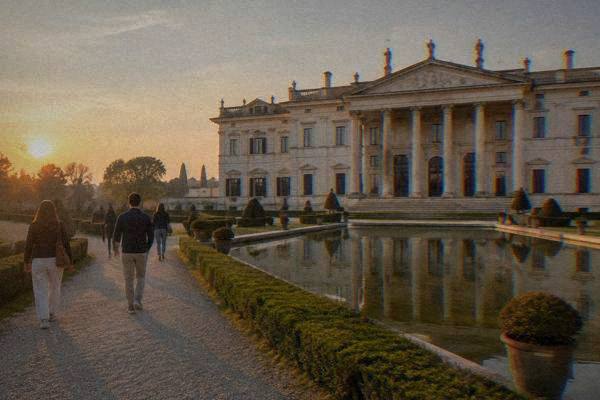 A few visitors seen from behind on a gravel path before an immense pale Venetian villa with a long columned front and a still reflecting pool, Villa Pisani