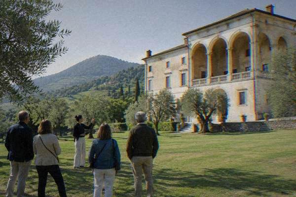 A guide and a small group on a lawn before a pale Renaissance villa with an open arched loggia on a green hillside, Villa dei Vescovi