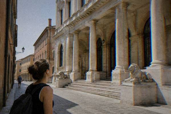 A lone visitor with earphones in, paused on a narrow street looking up at a pale neoclassical building with deep columned loggias, Caffe Pedrocchi