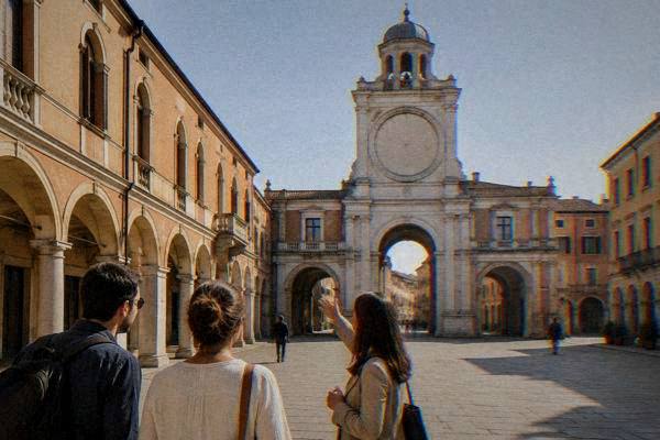 A private guide and two visitors in a paved square beneath a Renaissance clock tower with a tall central archway, Torre dell'Orologio