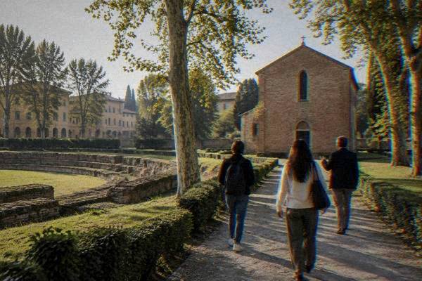 A private guide and two visitors seen from behind walking up a gravel path through a green garden toward a small plain red-brick chapel, Scrovegni Chapel