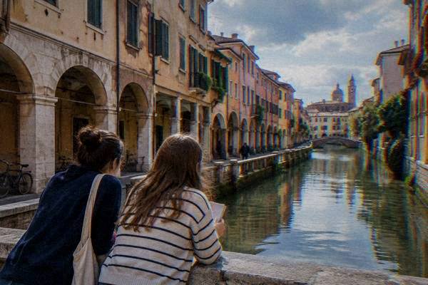 Two friends seen from behind leaning over a stone parapet on an arcaded street, one holding a small notebook, looking along the lane, Centro Storico