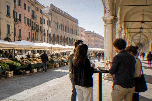 A small group sharing small plates round a high table on a pedestrian street under an arcade, glasses in hand, Piazza della Frutta
