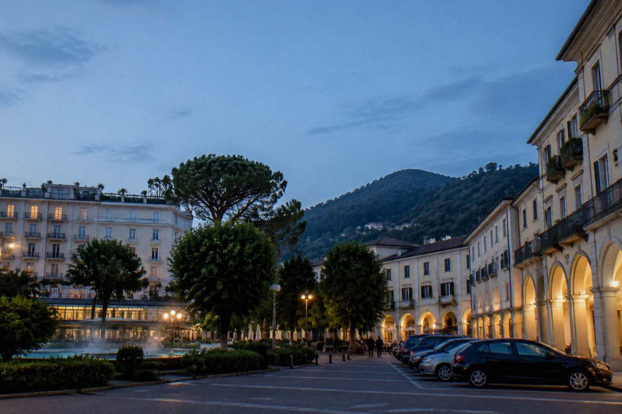 A thermal spa town square with parked cars, arcaded buildings and green hills rising behind