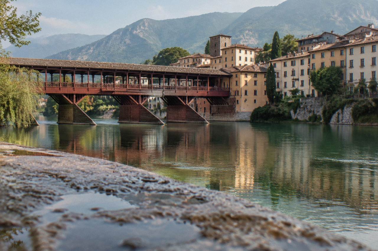A covered wooden bridge crossing a river with mountains rising behind a small Italian town