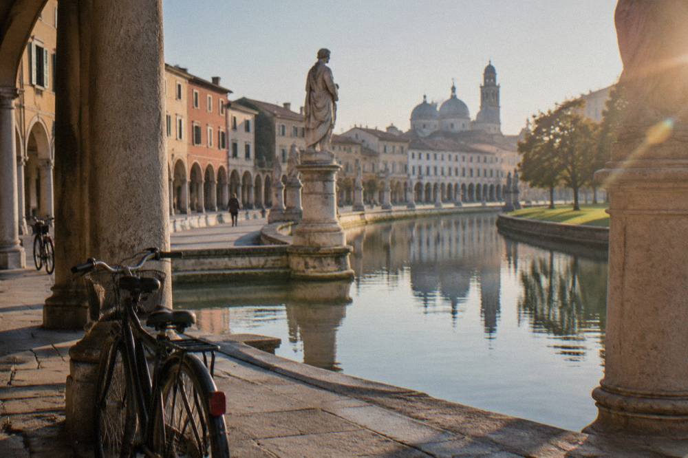 An elliptical square in Padua with a canal, ringed by statues and green lawns under a clear sky