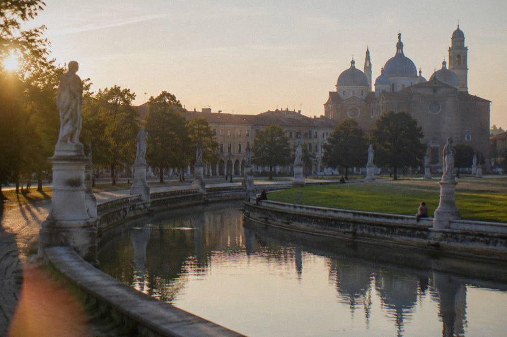 A canal lined with stone statues curving around the grassy island of Prato della Valle in Padua