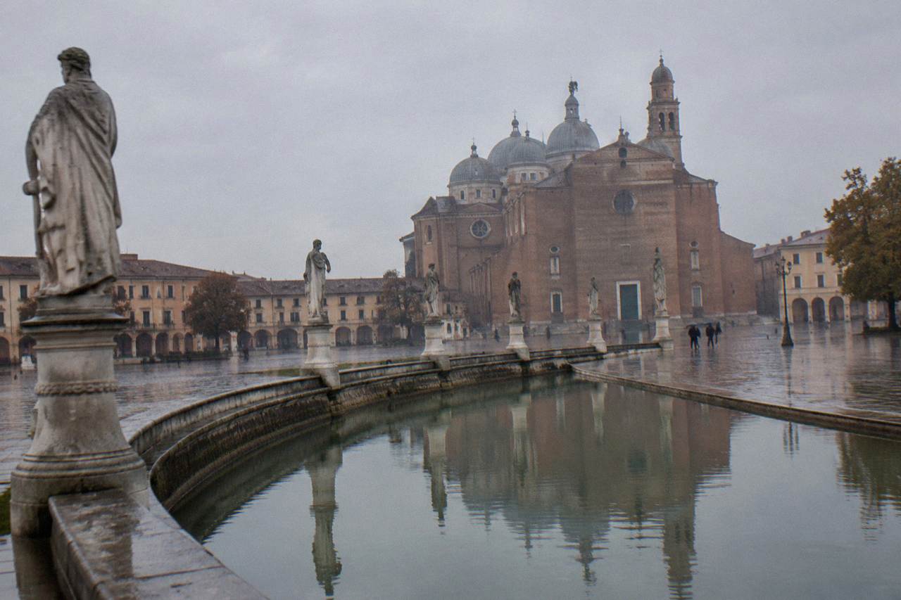 A large domed abbey church seen across a wide square ringed by a canal and statues