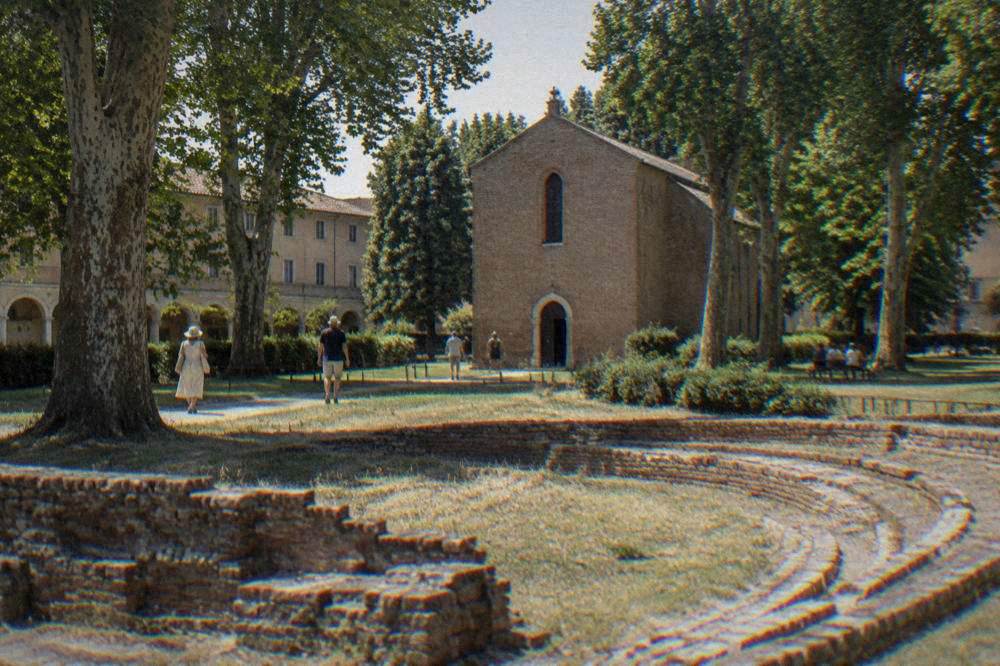 Plain brick chapel set in a green garden beside the remains of a Roman arena, morning light