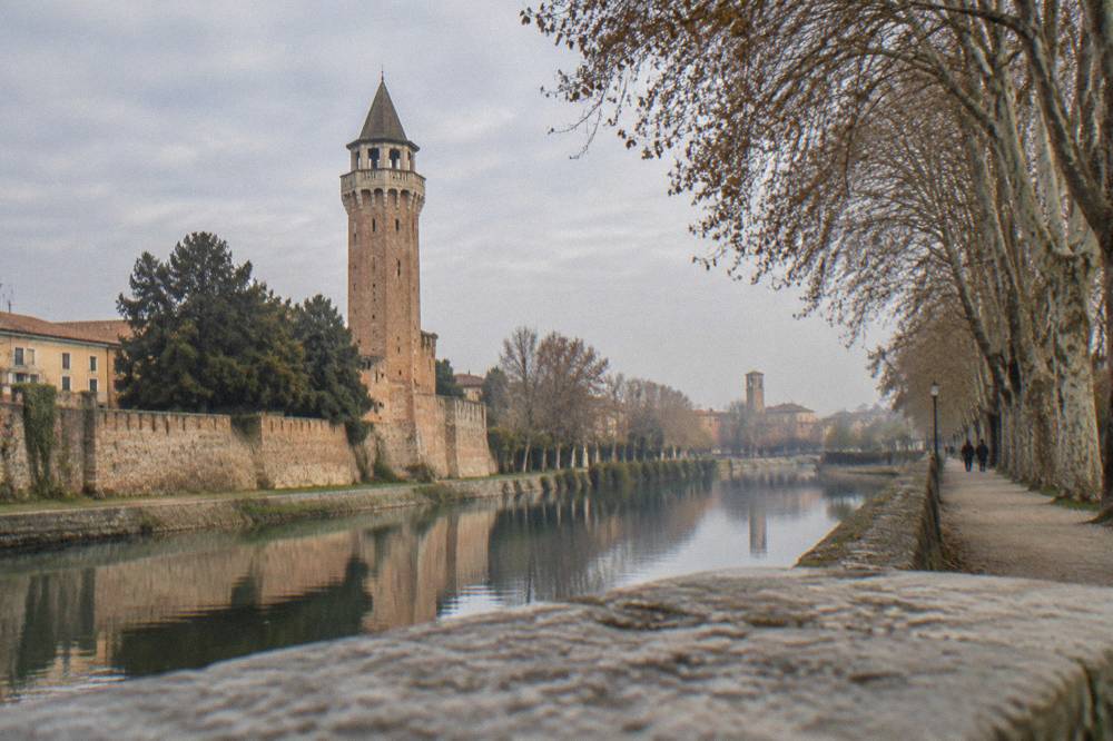 A tree-lined riverside path along an old city wall with a tower visible in the distance