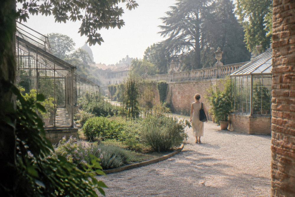A visitor walking a gravel path between greenhouses and old brick walls in a university garden