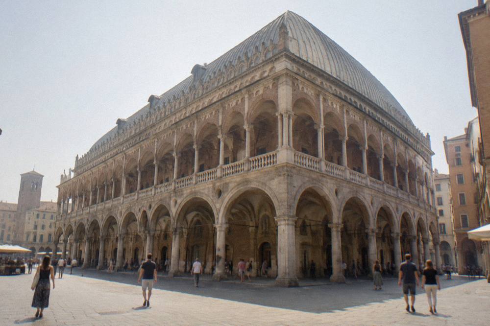 People walking under the stone arcades beside the Palazzo della Ragione on a bright day