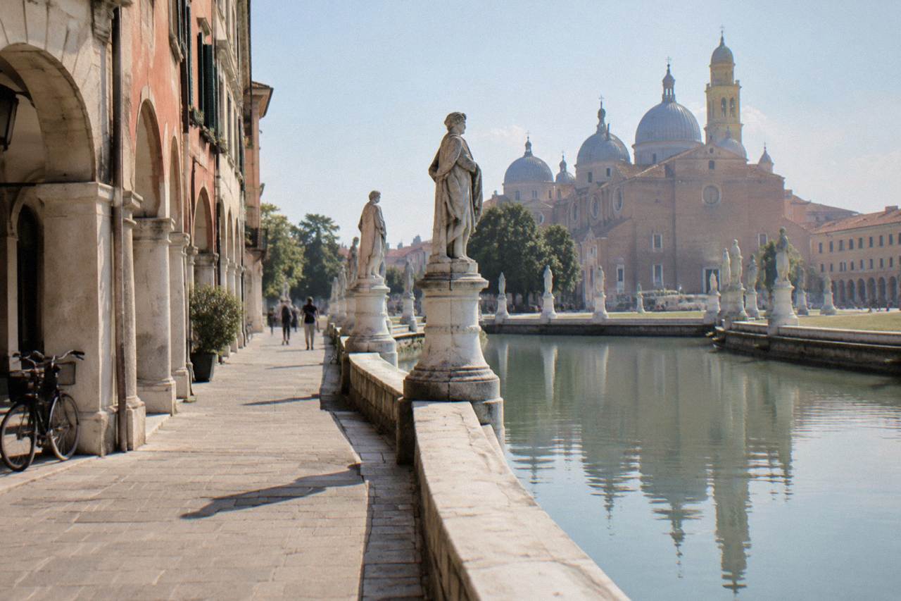 A wide oval square in Padua with a canal ringed by statues and a church dome in the background
