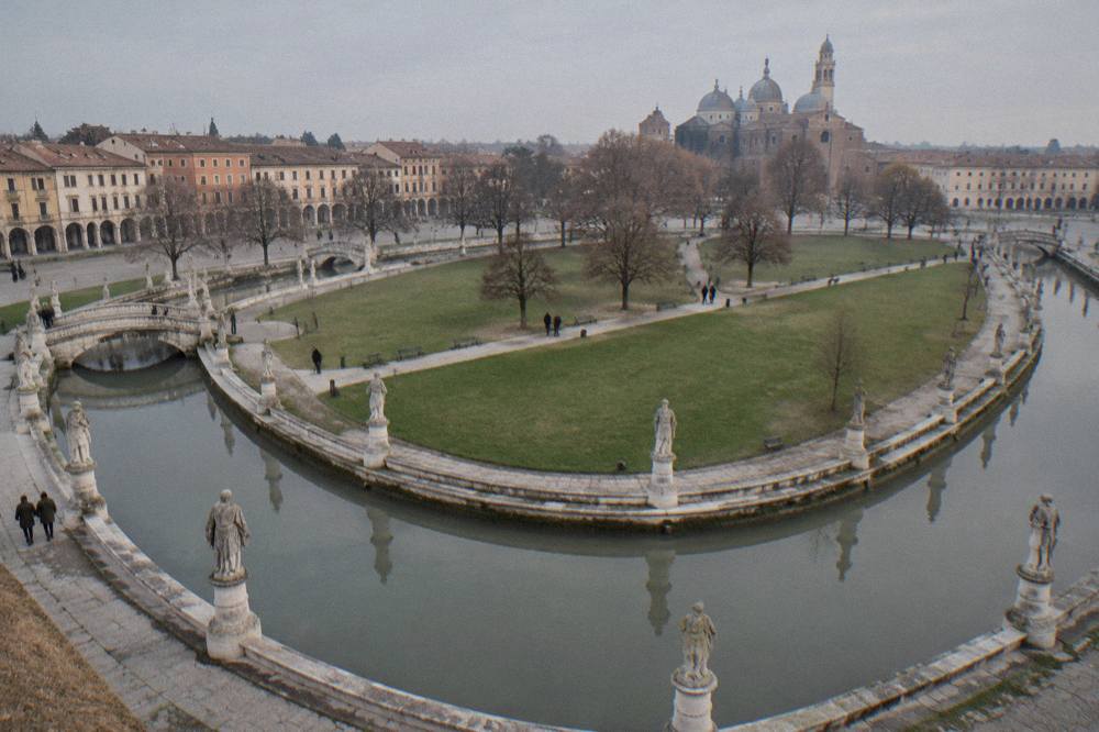 Statues on pedestals along a canal in an oval square, with bridges crossing to a grassed island