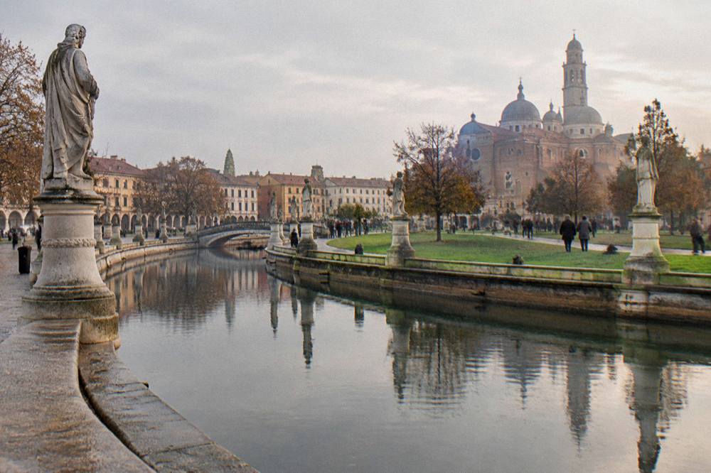 People strolling on a grassed island inside a canal ring, with bridges and statues around