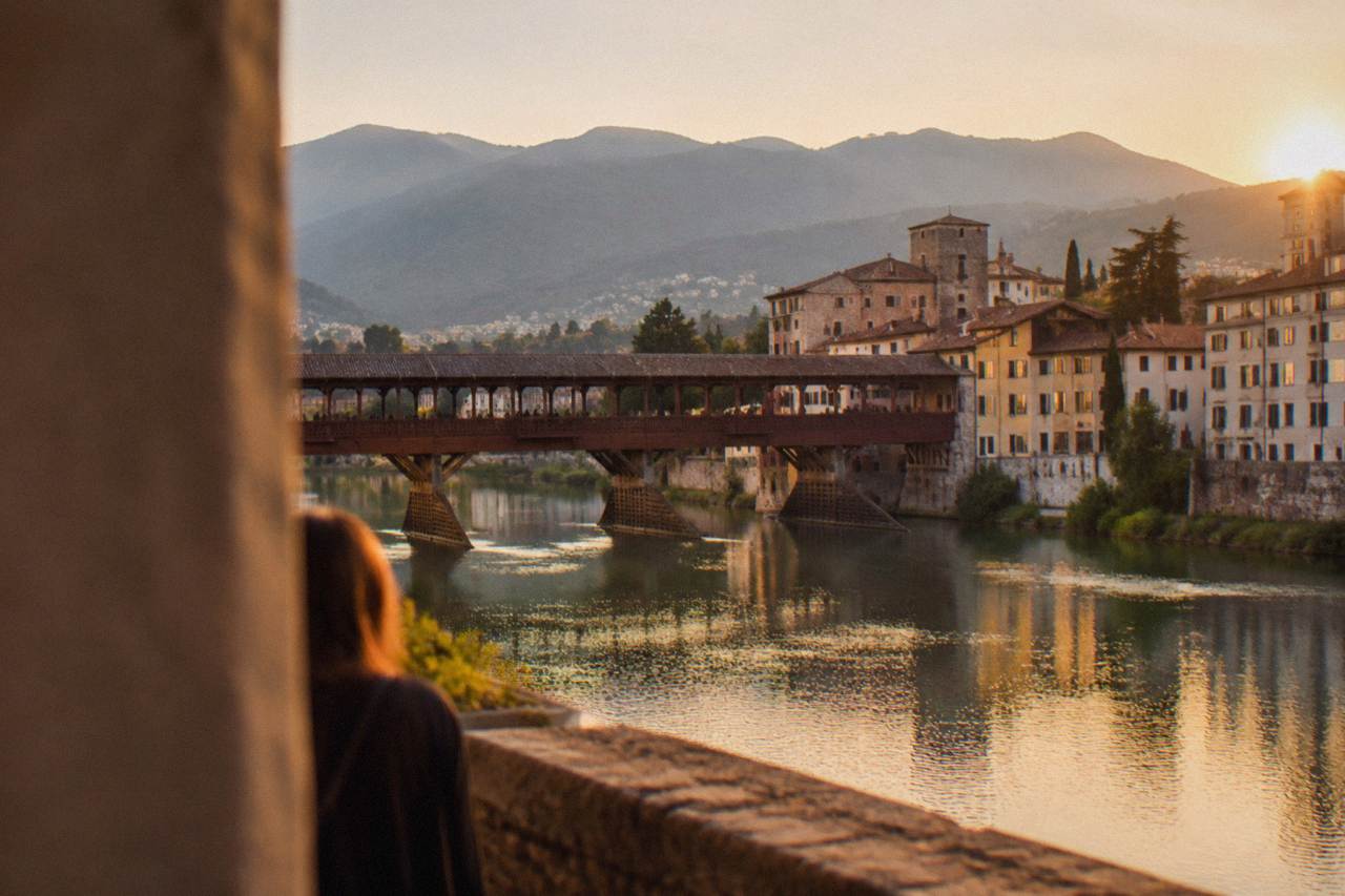 A covered wooden bridge crossing a river with mountains rising behind a small Italian town