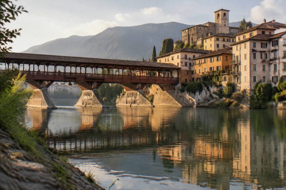 A roofed wooden bridge reflected in a green river at the edge of an old Italian town