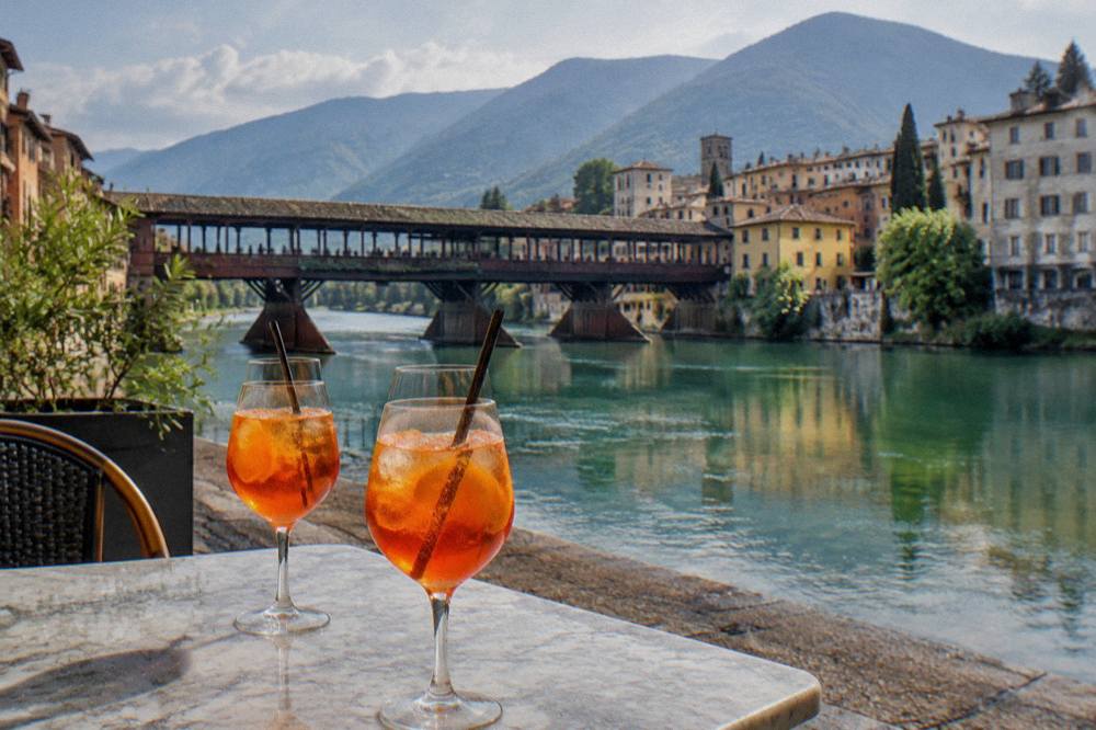 Two orange spritz cocktails on a marble table in a sunlit Italian square