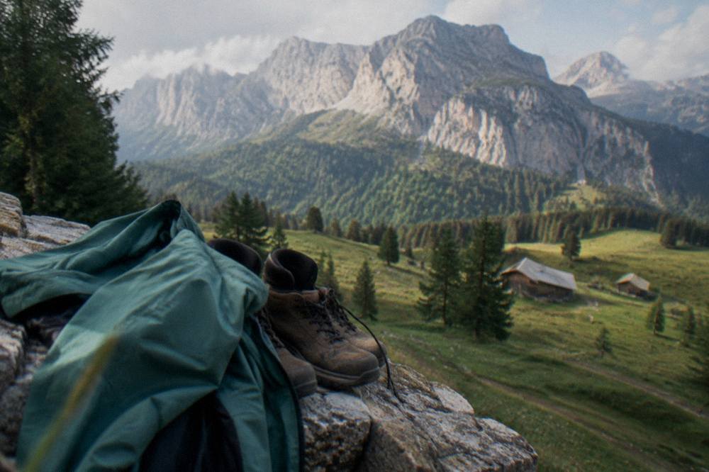 A folded rain jacket and hiking boots resting on a stone wall with mountains behind