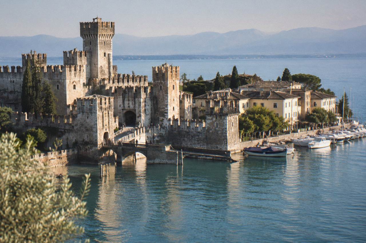 A lakeside peninsula town on Lake Garda with a stone fortress at the water's edge and boats moored along the shore