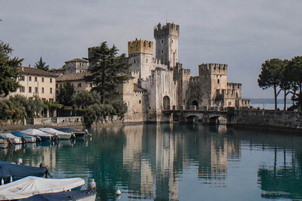 A medieval castle with towers reflected in a lake beside a small harbour of pleasure boats