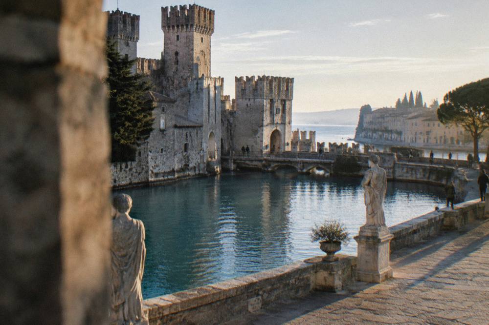 A wide stone square with a canal, ringed by statues, at golden hour in an Italian city