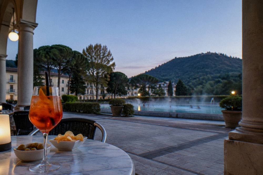 A glass of orange spritz and small snacks on a marble cafe table in a porticoed square