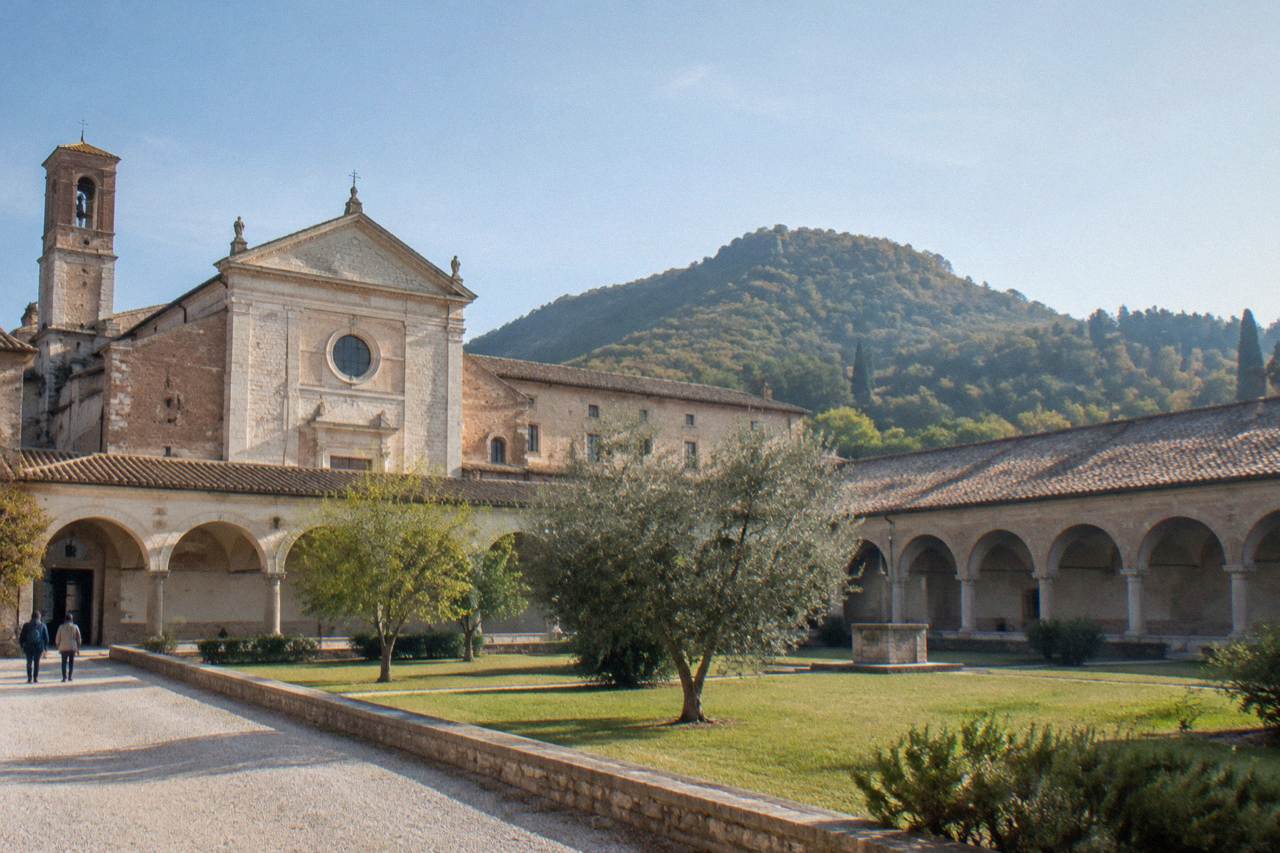 A stone Benedictine abbey building with a cloister courtyard and hills behind it