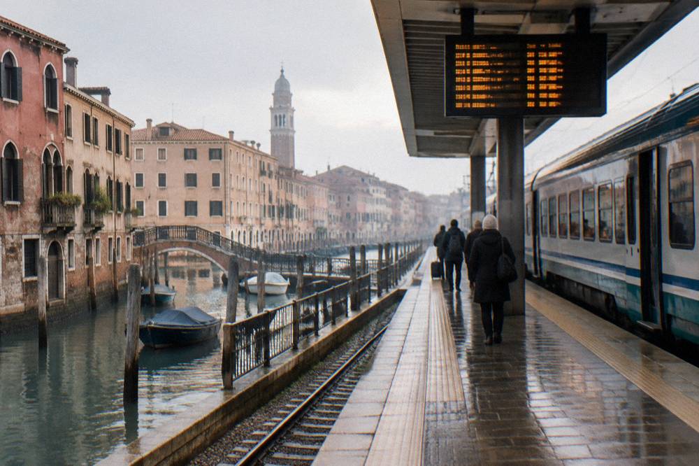 A regional train standing at a bright Italian railway platform beside a departures board