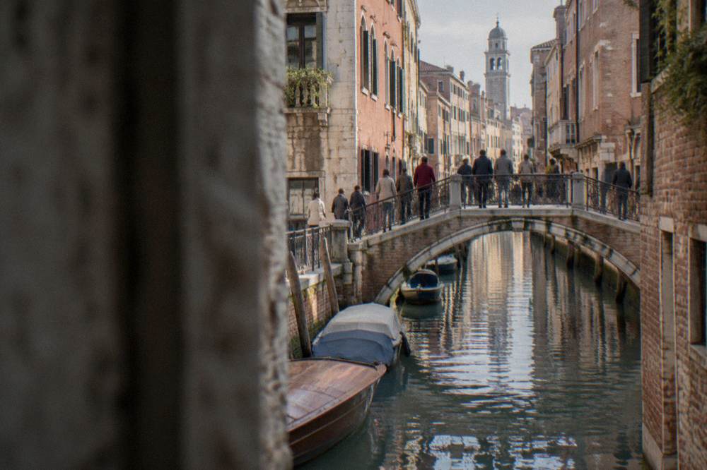 Crowds crossing a small arched bridge over a narrow canal lined with brick buildings