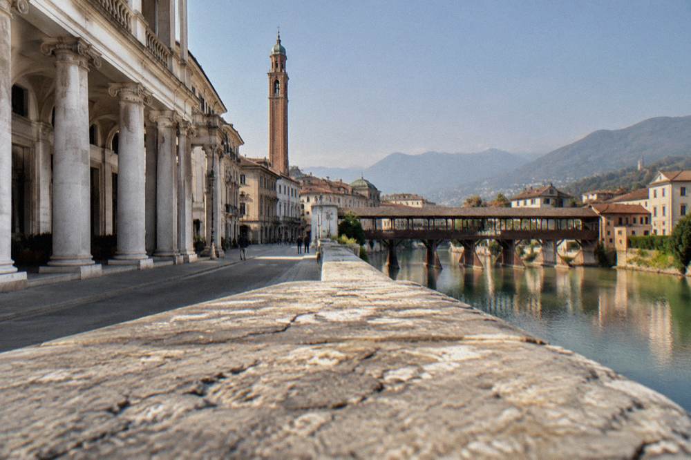 A covered wooden bridge over a river with mountains behind a small Italian town