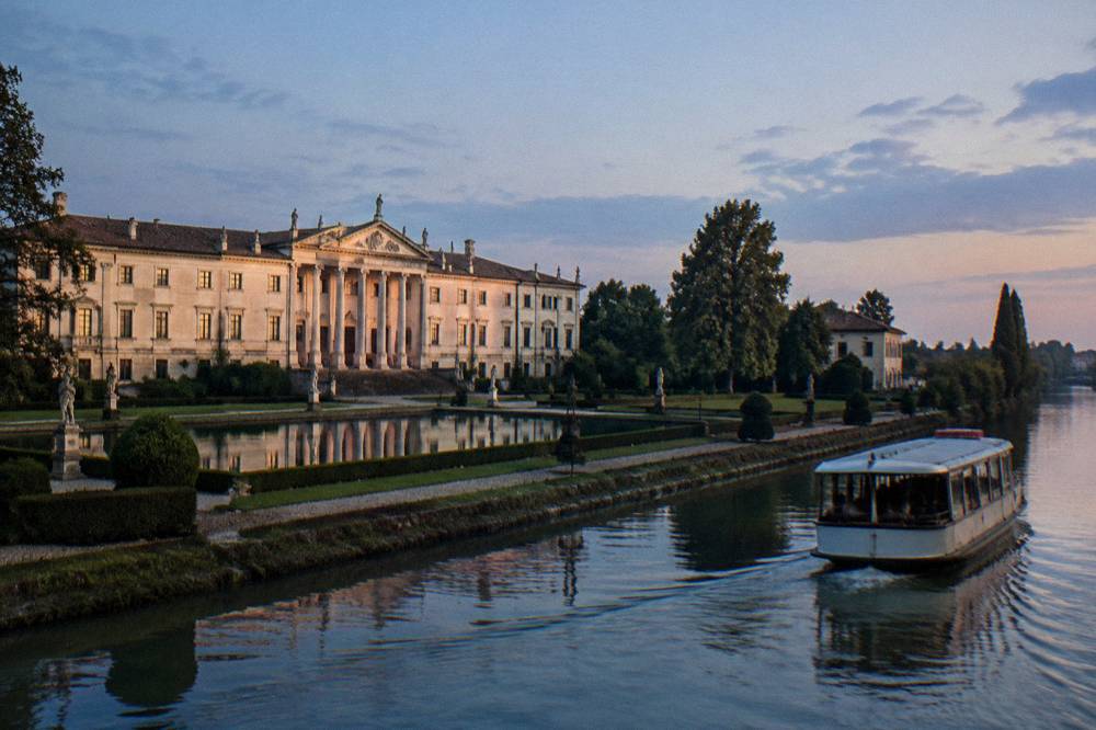 A passenger boat cruising along a canal lined with trees and villas
