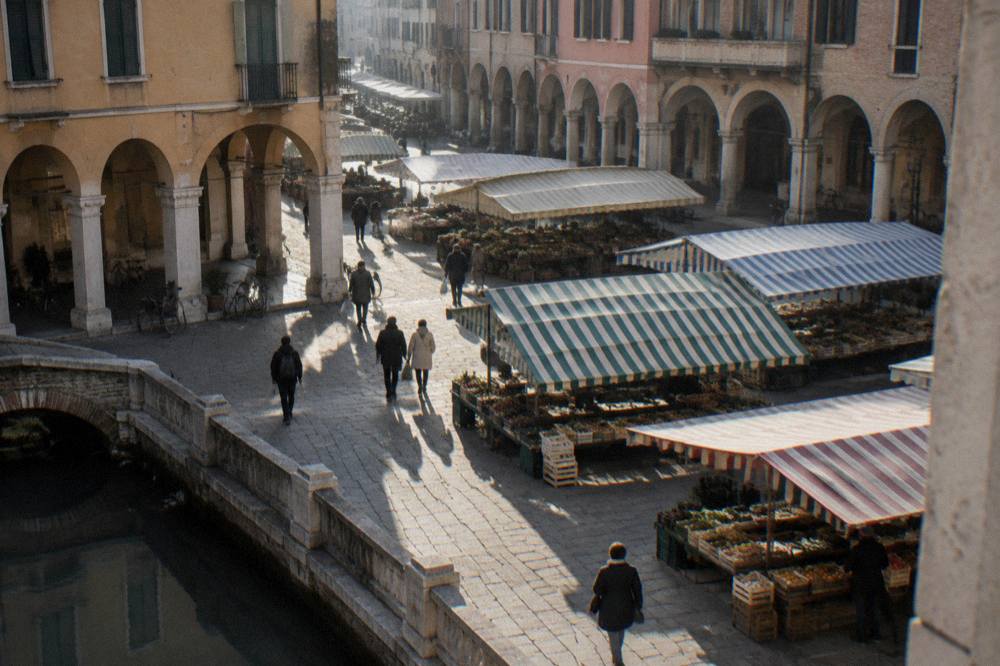 A sunlit piazza in Padua with market stalls shaded by striped awnings and arcaded buildings behind
