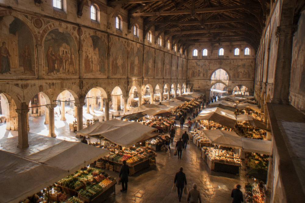A large medieval market hall in Padua with a soaring wooden ceiling and frescoed walls above stalls