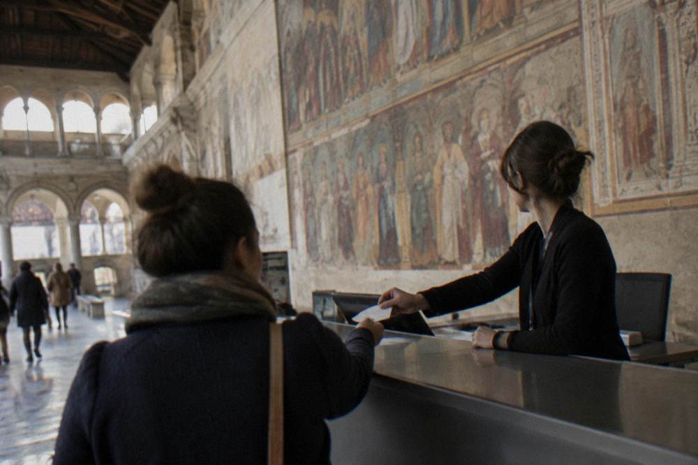 A clerk handing a timed-entry ticket to a visitor at a museum counter in front of a frescoed wall