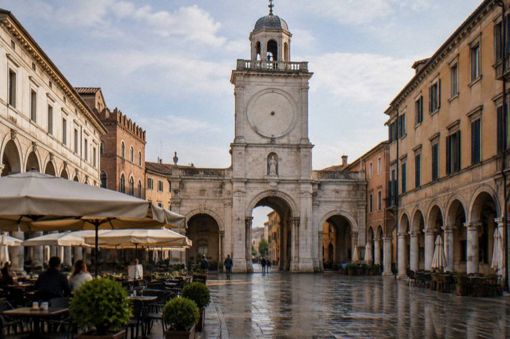 A historic clock tower rising above a pale Italian piazza with cafe tables below
