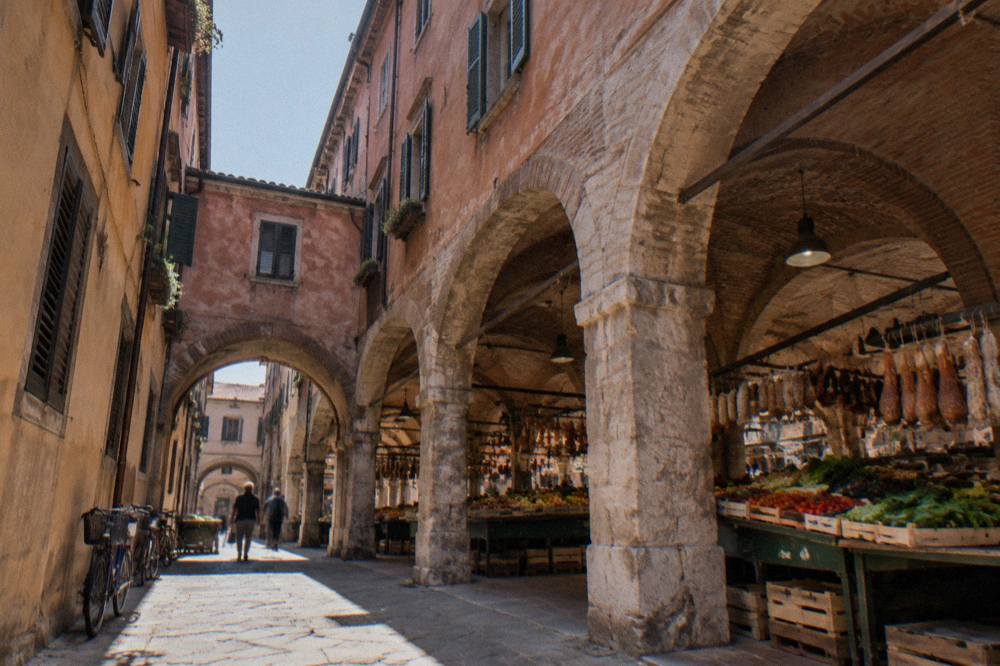 A covered market hall with produce stalls under stone arches and hanging cured meats