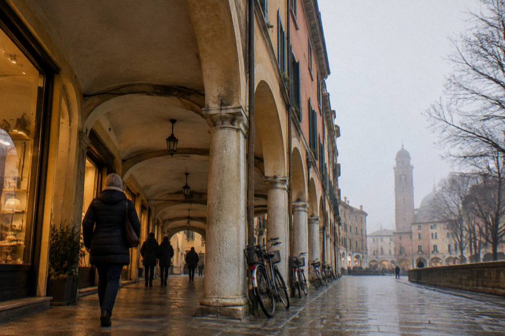 A traveller in a warm coat walking under the arcades of Padua past lit shop windows